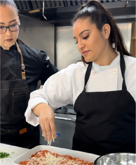 Chef apprentice sprinkles cheese over dish while mentor watches.