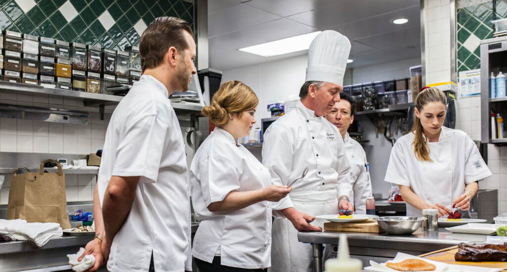 A group of students surrounding a mentor in a kitchen to illustrate how to become a chef.