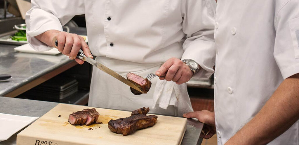 Two chefs at a cutting board, one of which is cutting a piece of meat to illustrate what are the duties of a chef